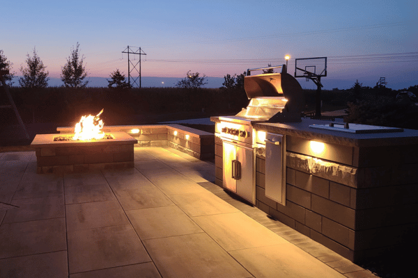 Landscape lighting illuminates outdoor grilling area, with a fire feature in the background.